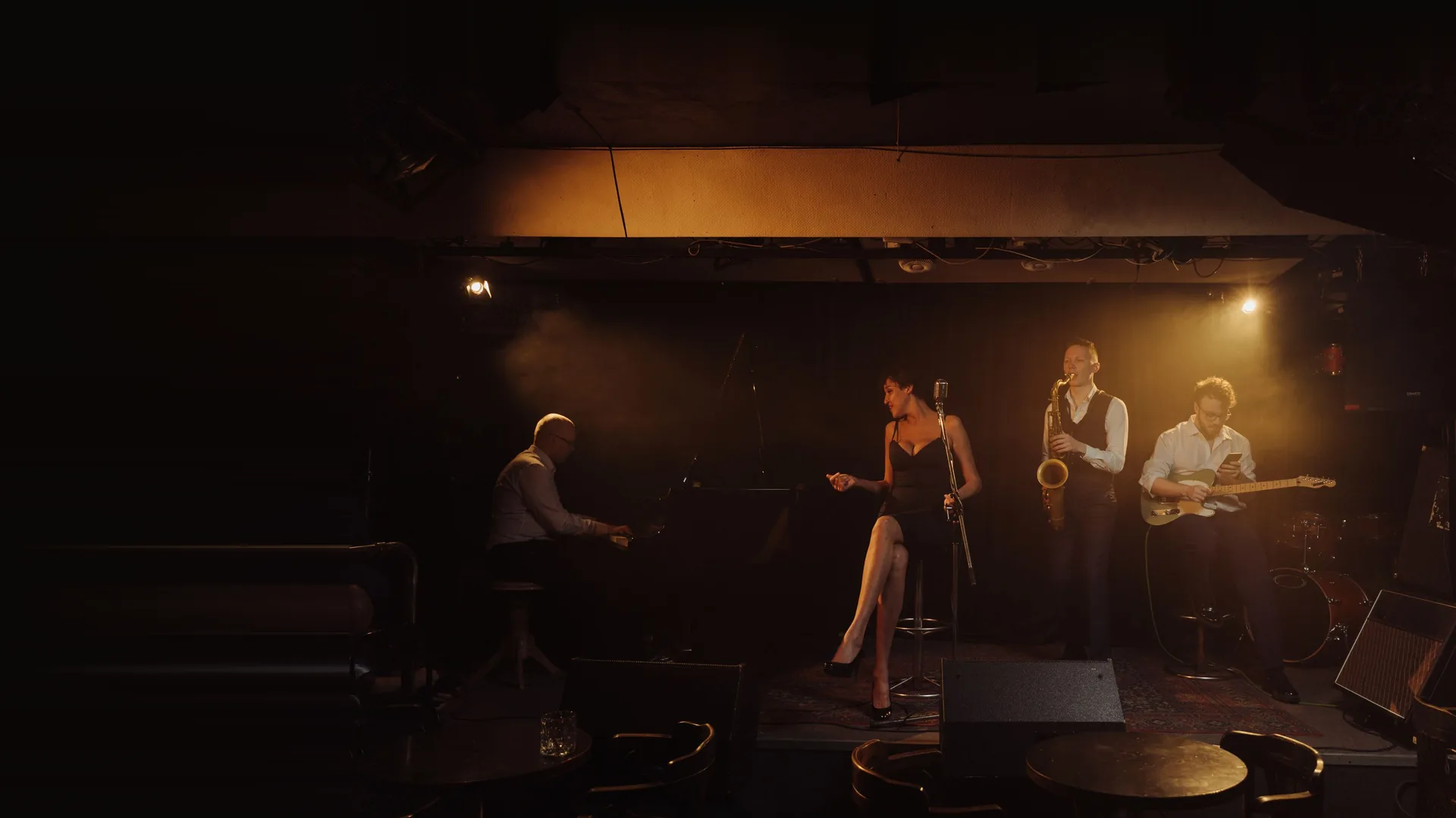 A jazz quartet on a small, warmly lit club stage: a pianist at a grand piano, a seated singer at a vintage microphone, a saxophonist and a guitarist, with a dark audience area in the foreground.