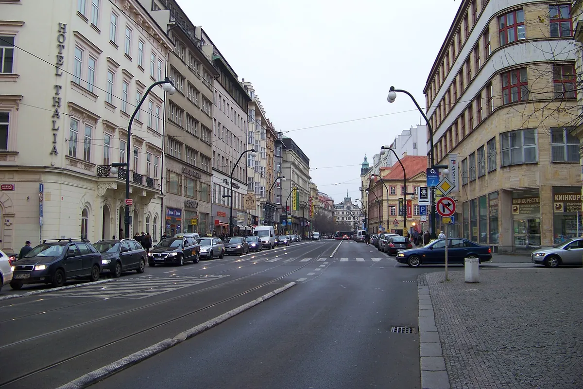 A wide Prague street with tram tracks and parked cars, lined with tall cream and stone buildings, running away toward the distance.