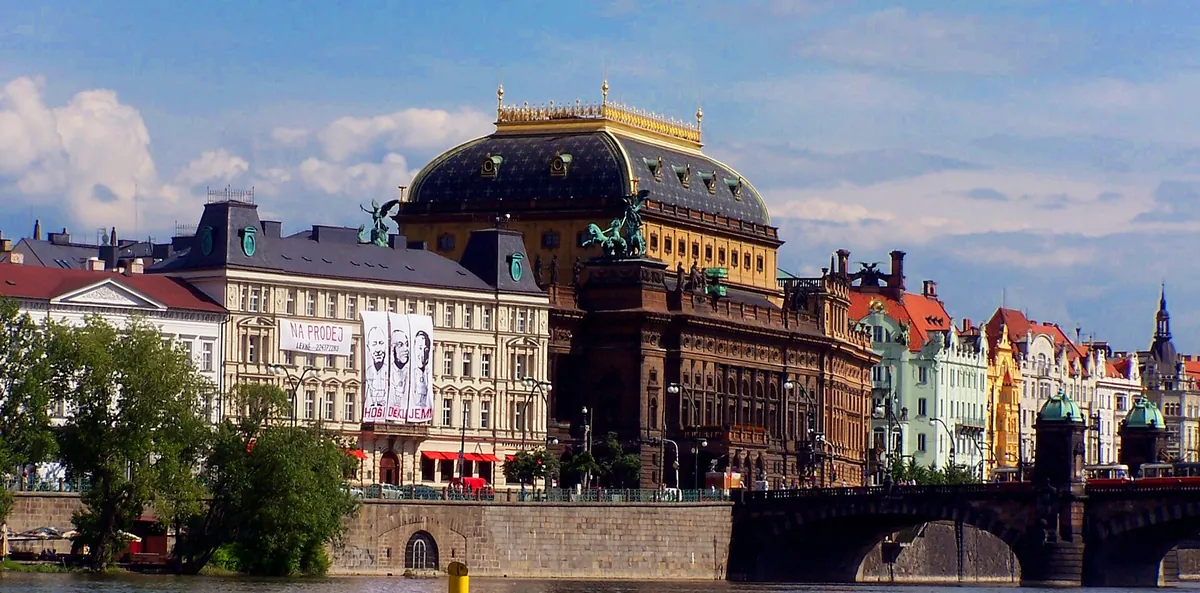 The National Theatre in Prague seen across the Vltava, with its dark dome crowned in gold beside the Legion Bridge.
