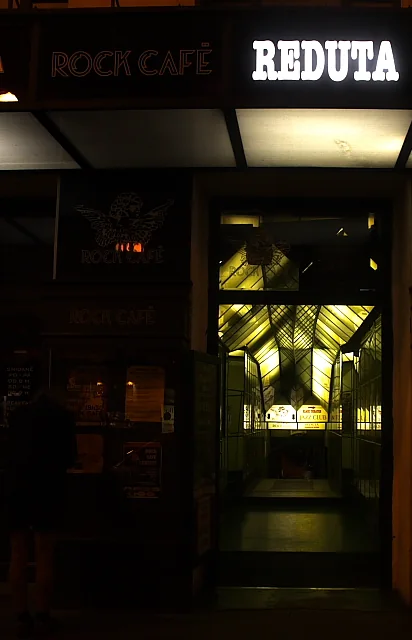 A lit doorway on a Prague street at night, with a large illuminated REDUTA sign above it and a glass-roofed passage leading inside.