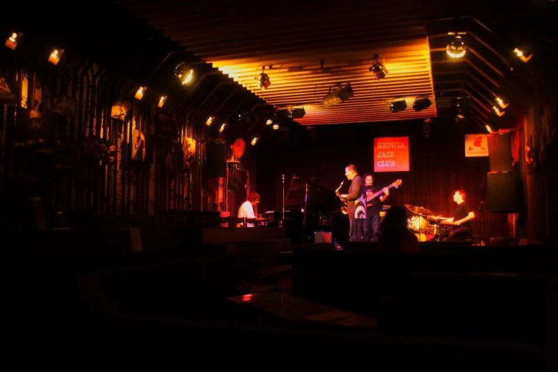 A jazz band playing on a small stage lit in orange, with a red REDUTA JAZZ CLUB sign behind the musicians and a dark audience area in front.